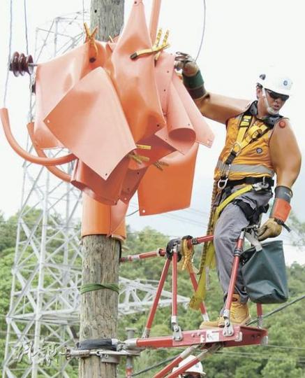圖中的技工正準備更換電線桿上的菲士(fuse)座,技工除戴頭盔,還需要站在絕緣工作台(紅色架)、戴上防衝擊太陽眼鏡、絕緣臂袖和絕緣手套,而電線桿亦要鋪滿絕緣布,避免觸電。(歐文瀚攝)站在絕緣升降工作台上的譚昭義示範用絕緣棍協助更換架空電纜終端頭。(歐文瀚攝) 圖中的技工正準備更換電線桿上的菲士(fuse)座,技工除戴頭盔,還需要站在絕緣工作台(紅色架)、戴上防衝擊太陽眼鏡、絕緣臂袖和絕緣手套,而電線桿亦要鋪滿絕緣布,避免觸電。(歐文瀚攝)站在絕緣升降工作台上的譚昭義示範用絕緣棍協助更換架空電纜終端頭。(歐文瀚攝)
