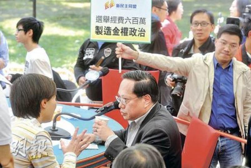 民主黨主席何俊仁（中）昨日出席《城市論壇》時，黨友、但已另起爐灶成立新民主同盟的范國威亦到場舉牌，抗議政改方案選舉經費過高。圖左一背向鏡頭者為政制及內地事務局副局長黃靜文。（尹錦恩攝）