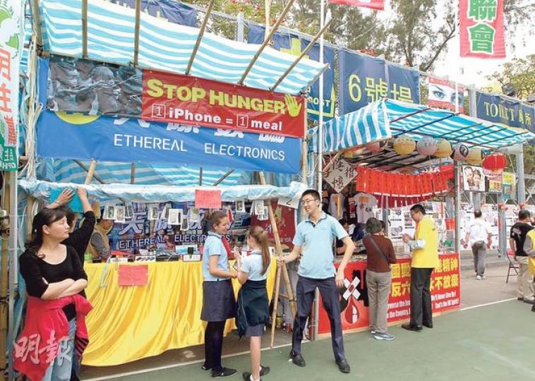 維園2號攤檔(左方)現由國際學校售賣手機套作慈善籌款,前天掛在檔口的「天際數碼」橫額,被另一寫上英文標語(停止飢餓 1個iPhone=1餐飯)及饑民照片的橫額蓋着。攤檔昨日有數名穿便服成年男女(包括左一黑衣女子)看檔,3名身穿國際學校校服的學生(藍衣者)則在檔前宣傳拉客。右為支聯會的1號攤位。(李澤彤攝)支聯會今年在維園只有一攤檔,繼續主打常委張文光(左三)等人寫揮春,主席李卓人(左二)期望籌得230萬元,以興建六四永久紀念館。(李澤彤攝) 維園2號攤檔(左方)現由國際學校售賣手機套作慈善籌款,前天掛在檔口的「天際數碼」橫額,被另一寫上英文標語(停止飢餓 1個iPhone=1餐飯)及饑民照片的橫額蓋着。攤檔昨日有數名穿便服成年男女(包括左一黑衣女子)看檔,3名身穿國際學校校服的學生(藍衣者)則在檔前宣傳拉客。右為支聯會的1號攤位。(李澤彤攝)支聯會今年在維園只有一攤檔,繼續主打常委張文光(左三)等人寫揮春,主席李卓人(左二)期望籌得230萬元,以興建六四永久紀念館。(李澤彤攝)