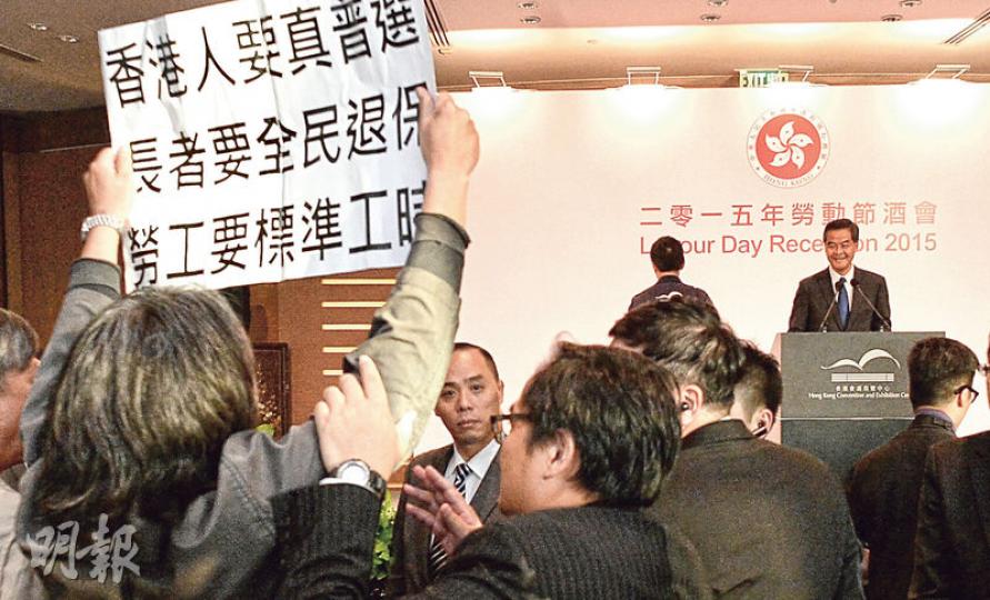 社民連立法會議員梁國雄(左一背鏡頭者)噚日去勞動節酒會示威,佢被趕走時台上嘅特首梁振英忍唔住笑。(葉家豪攝) 社民連立法會議員梁國雄(左一背鏡頭者)噚日去勞動節酒會示威,佢被趕走時台上嘅特首梁振英忍唔住笑。(葉家豪攝)